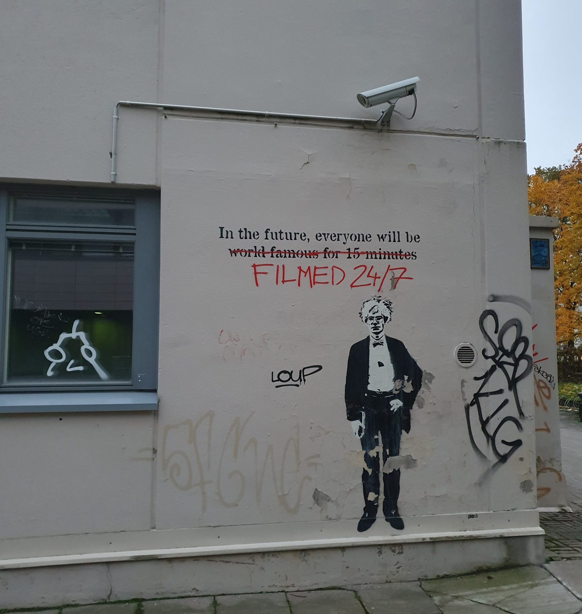Graffiti Itä-Pasilassa. Andy Warholin kuva, sekä lainaus "In the future, everyone will be" ja sen jälkeen yliviivattuna "world famous for 15 minutes" ja tilalle kirjoitettuna "filmed 24/7". Graffitin yläpuolella seinässä on valvontakamera.