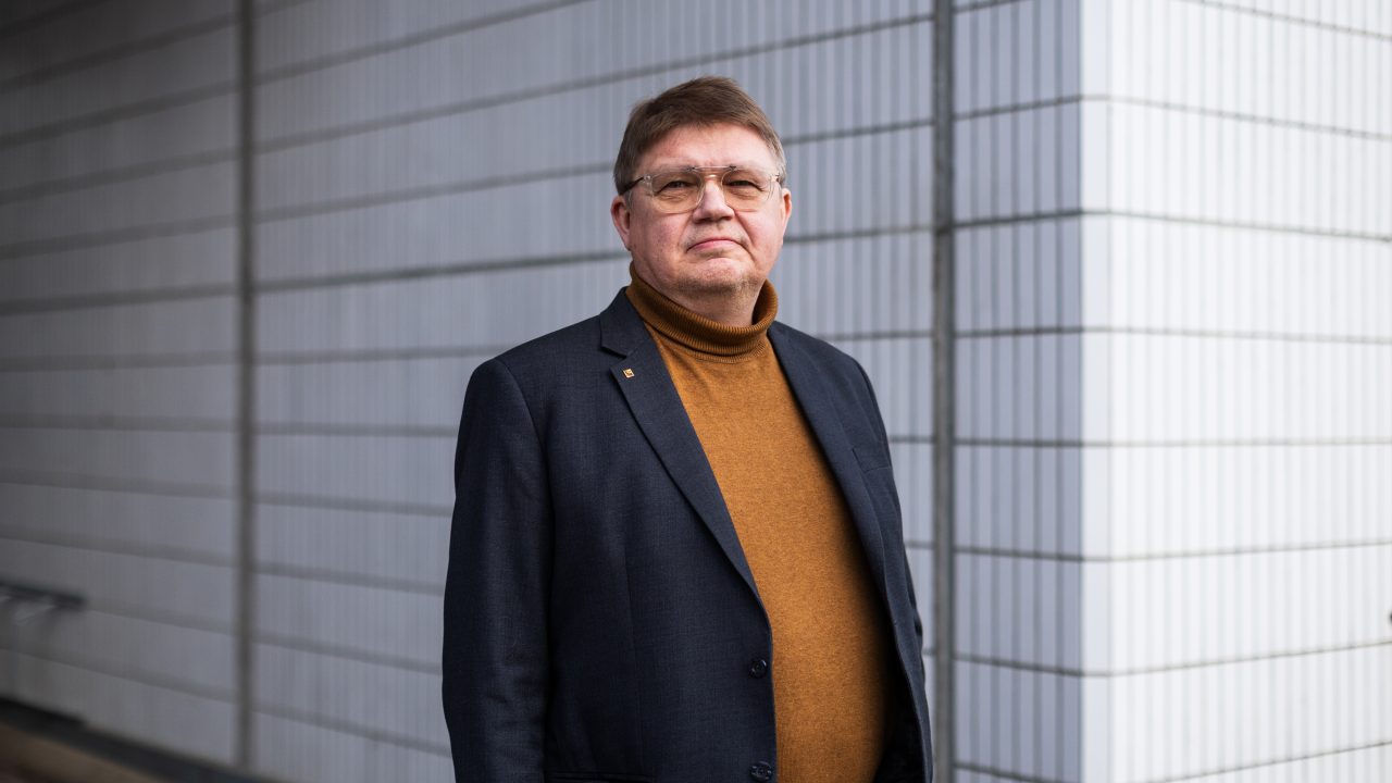 Pikkutakkiin ja kellanruskeaan poolopaitaan pukeutunut mies katsoo puolivakavana kohti. Taustalla kerrostalon vaalea kaakeliseinä.